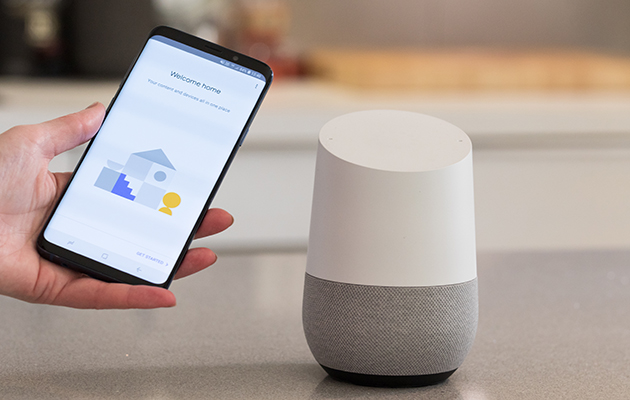 Styr din smarta belysning med hjälp av din röst En hand håller en smartphone intill en Google Home smarthögtalare.