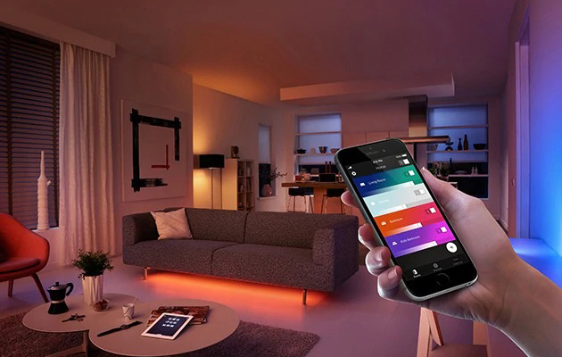Synka din belsyning med soluppång och solnedgång En hand som håller i en smartphone framför ett färgglatt upplyst vardagsrum.