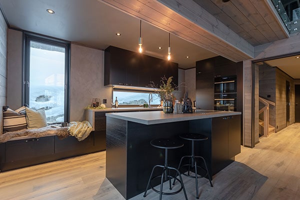 Översiktsbild av köket med mörka fronter och grå betongbänkskiva, bänk framför fönstret.