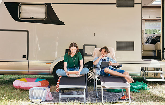 Underhållning på semestern Två ungdomar, en pojke och en flicka som sitter i campingstolar framför en husbil och använder en surfplatta