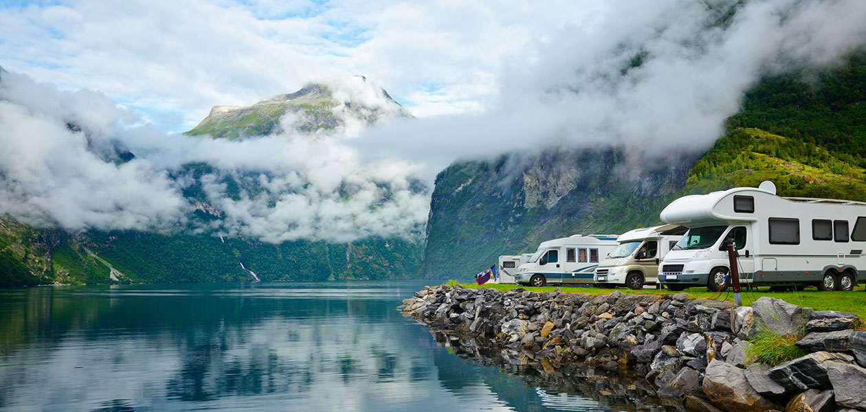 Bekväm campingsemester En rad av vita husbilar som står längs vattnet med fjäll och moln i bakgrunden