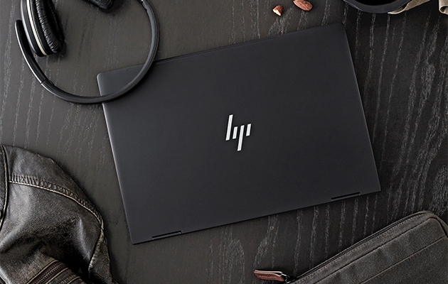 Datorn HP Envy x360 på ett svart bord med hörlurar, en väska och jacka bredvid sig” title=
