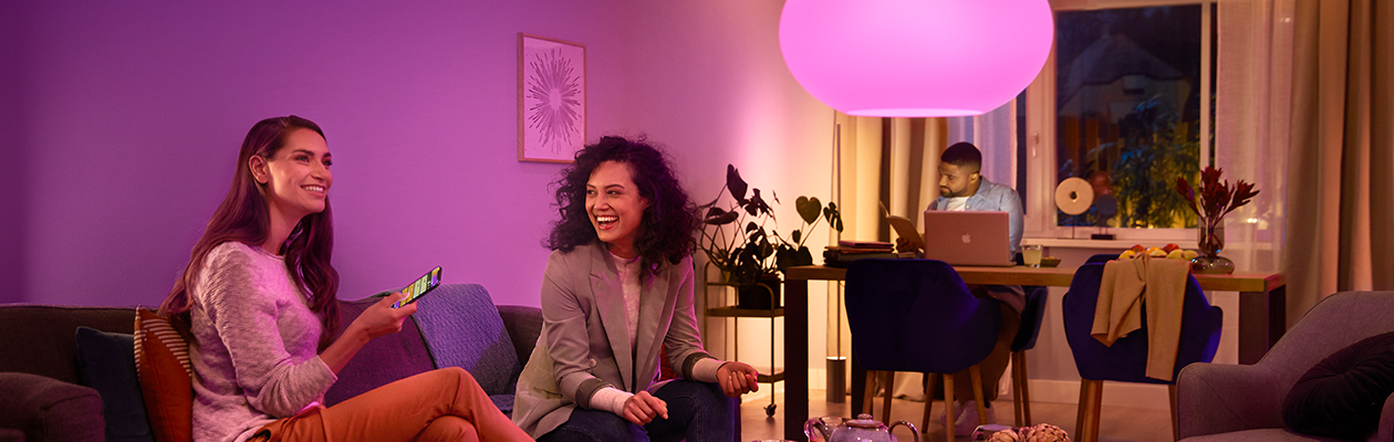 Philips Hue – Bridge vs Bluetooth Två kvinnor som sitter i en soffa och en man som sitter vid ett bord i bakgrunden. Philips Hue lampa med lila ljus i taket.
