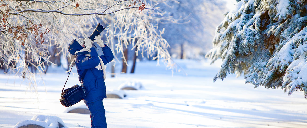Månadens fotodeal En fotograf klädd i blå vadderade vinterkläder som fotograferar rimfrost på en gren ovanför dem, snöig t landskap i bakgrunden