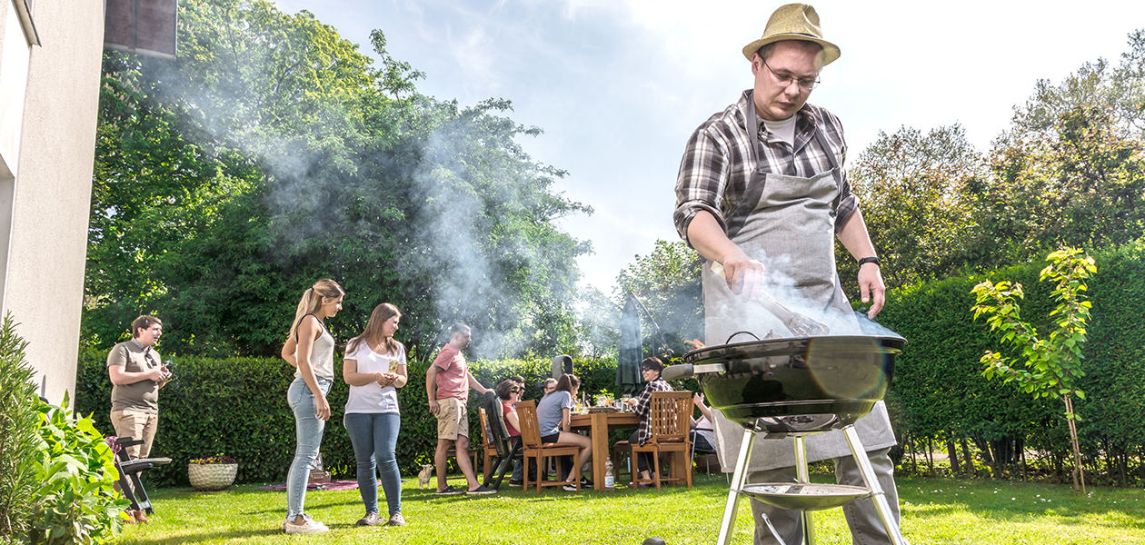 Grilltillbehör En man som grillar i förgrunden och en grupp av människor sitter och står i bakgrunden