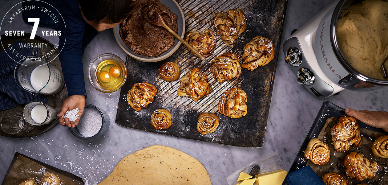 Ankarsrum – din vän i köket Ankarsrum köksmaskin på en köksbänk, kanelbullar, ett barn och en logotyp med 7-års garanti