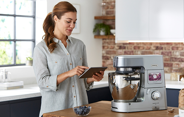 Kenwood World-appen En kvinna som använder en surfplatta och står intill en Kenwood köksmaskin
