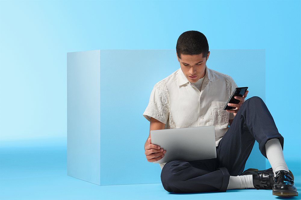 Windows 11 låter dig maximera kreativiteten Man sitter på golvet med en Windows 11-dator i famnen och en mobil i handen, på en blå bakgrund