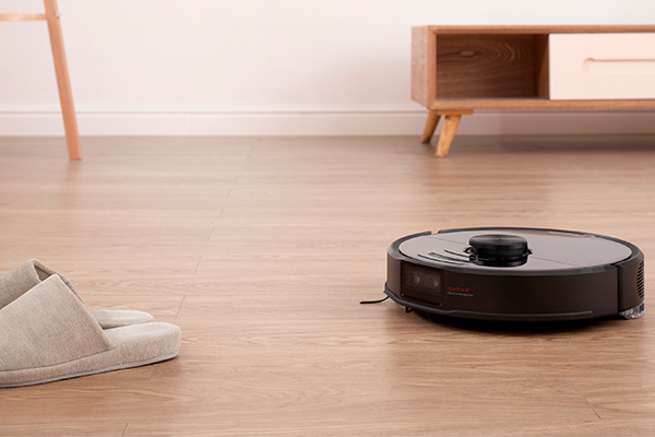 Roborock En svart robotdammsugare som dammsuger ett golv med ett par tofflor bredvid