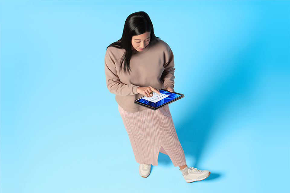 Få ett nytt perspektiv Kvinna i beiga kläder står med Windows 11 2-i-1 I händerna framför blå bakgrund