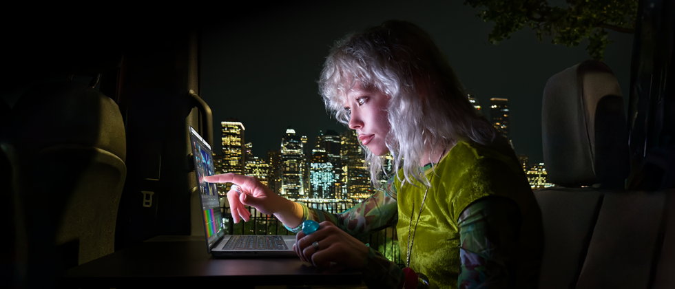 En person sitter i baksätet av en skåpbil, genom den öppna passagerardörren skymtar stadens siluett på kvällen, en MacBook Pro med PROLED XDR-skärm står på ett skrivbord, personen är upplyst av skärmen och använder touch-funktionen för att redigera en video i DaVinci Resolve Studio
