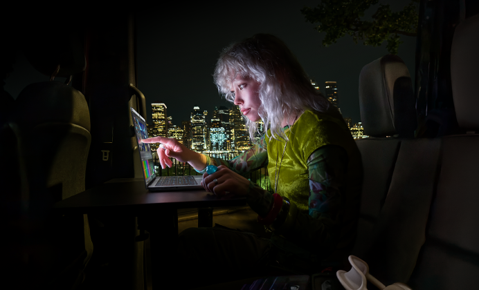 En person sitter i baksätet av en skåpbil, genom den öppna passagerardörren skymtar stadens siluett på kvällen, en MacBook Pro med PROLED XDR-skärm står på ett skrivbord, personen är upplyst av skärmen och använder touch-funktionen för att redigera en video i DaVinci Resolve Studio