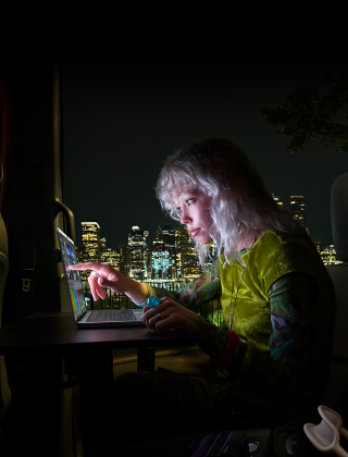 En person sitter i baksätet av en skåpbil, genom den öppna passagerardörren skymtar stadens siluett på kvällen, en MacBook Pro med PROLED XDR-skärm står på ett skrivbord, personen är upplyst av skärmen och använder touch-funktionen för att redigera en video i DaVinci Resolve Studio