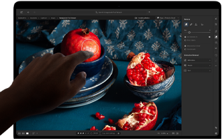 MacBook Pro med PROLED XDR-skärm som visar ett stilleben med granatäpplen samt blå keramikföremål och textilier, en hand sträcks ut mot skärmen för att interagera med bilden