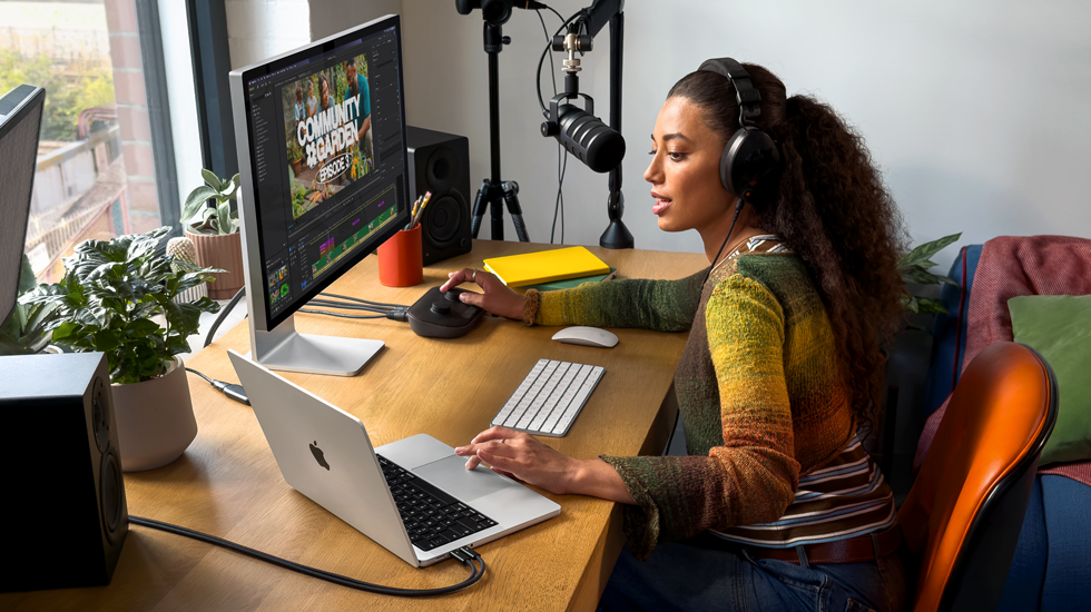 En kvinna med hörlurar sitter hemma vid sitt skrivbord och pratar i en mikrofon med MacBook Pro ansluten till en extern skärm
