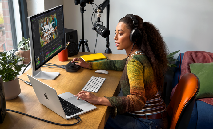 En kvinna med hörlurar sitter hemma vid sitt skrivbord och pratar i en mikrofon med MacBook Pro ansluten till en extern skärm