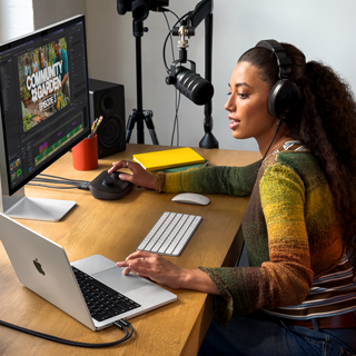 En kvinna med hörlurar sitter hemma vid sitt skrivbord och pratar i en mikrofon med MacBook Pro ansluten till en extern skärm