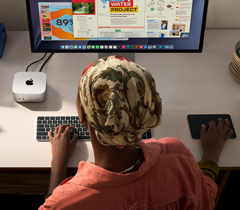 En person skriver vid ett skrivbord med en Mac mini med Studio Display ansluten, Magic Keyboard, Magic Trackpad och olika föremål