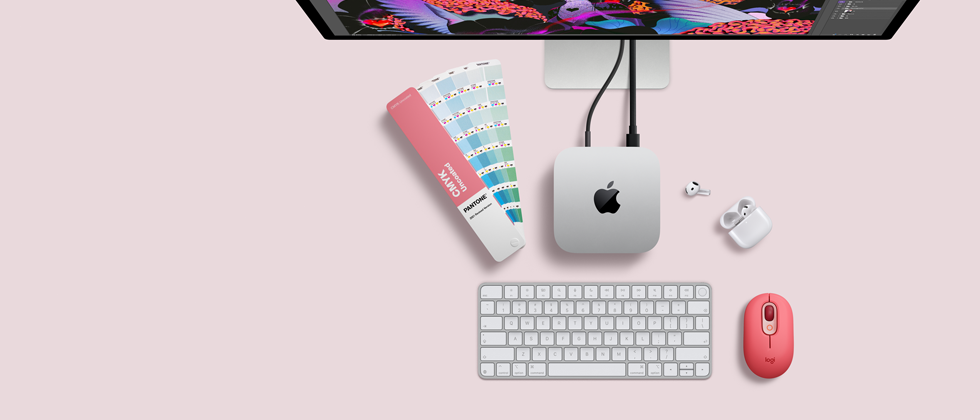 Mac mini sedd ovanifrån, silverfärgat aluminium, rundade hörn, blank svart Apple-logotyp i mitten, ansluten med en sladd, en del av Studio Display syns längst upp, öppna AirPods 4, Pantone-färgprover samt rött Logitech-tangentbord och röd mus