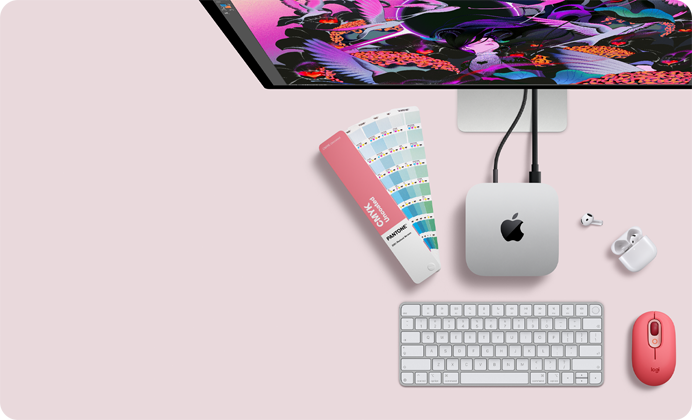 Mac mini sedd ovanifrån, silverfärgat aluminium, rundade hörn, blank svart Apple-logotyp i mitten, ansluten med en sladd, en del av Studio Display syns längst upp, öppna AirPods 4, Pantone-färgprover samt rött Logitech-tangentbord och röd mus
