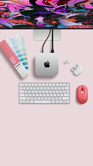 Mac mini sedd ovanifrån, silverfärgat aluminium, rundade hörn, blank svart Apple-logotyp i mitten, ansluten med en sladd, en del av Studio Display syns längst upp, öppna AirPods 4, Pantone-färgprover samt rött Logitech-tangentbord och röd mus