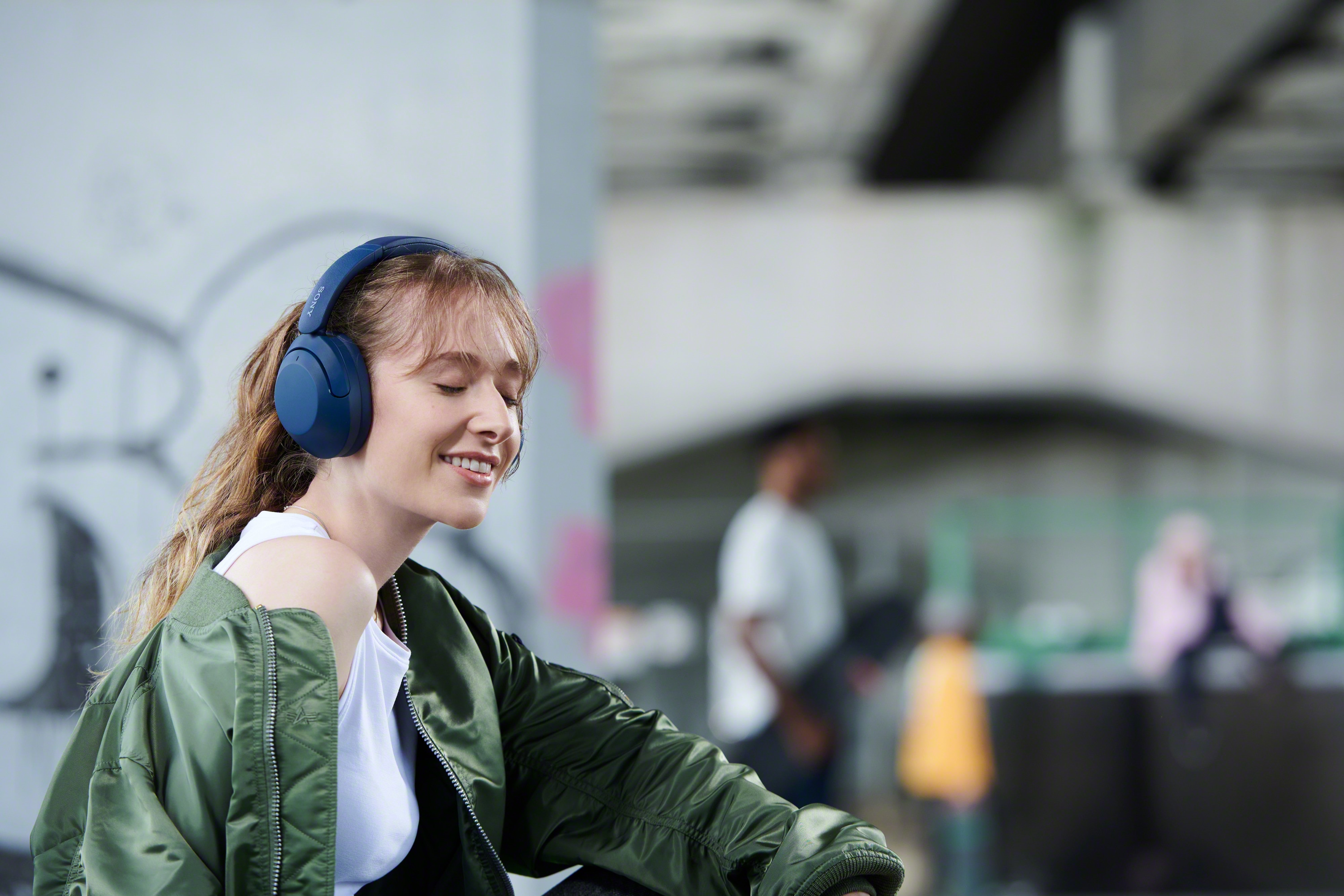 Glad kvinna med WH-XB910 hörlurar och grön jacka på en tunnelbaneperrong eller liknande