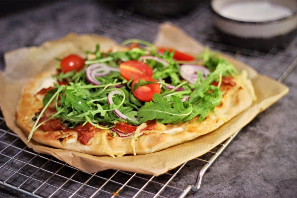 Hemmagjord pizza på bakplåtspapper med ruccolasallad, tomater, skinka och rödlök