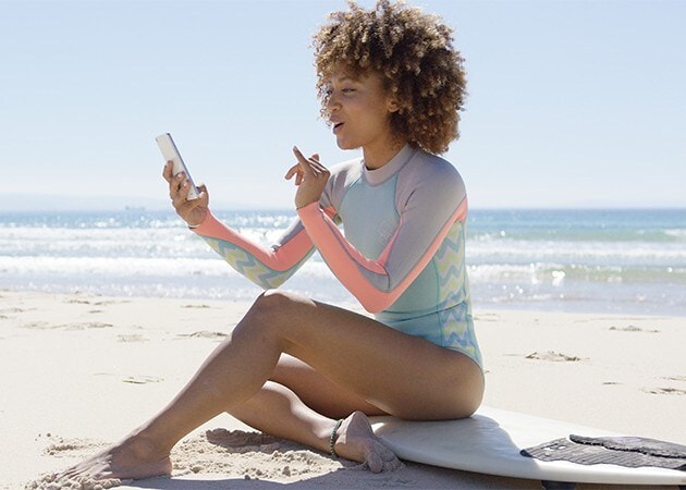 En kvinna sitter på en surfbräda på en strand med sin smartphone i handen.