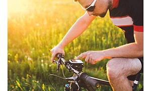 En man på en cykel med en GPS fäst på cykelstyret för guidning som han pekar på. Gult fält och solljus syns i bakgrunden. 