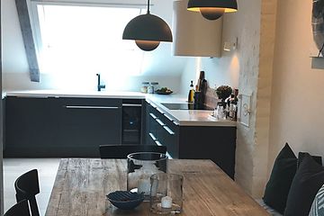 Köket sett från matbordet i trä, stilren design med grå färg och vit bänkskiva anpassat till lutande väggar och takbjälkar. . 