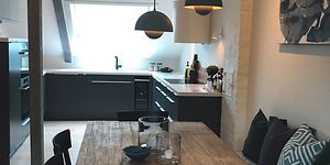 Köket sett från matbordet i trä, stilren design med grå färg och vit bänkskiva anpassat till lutande väggar och takbjälkar. .