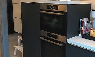 Två ugnar och kylskåp integrerade i köket som har mörkgrå färg. Ovanför är vita skåpshyllor för mer förvaring.
