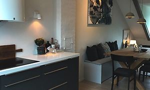 Ena hörnet av köket med induktionshäll, grå lådor och en vit bänkskiva. Matbord i trä med en grå soffa.