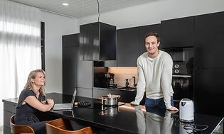 Ett par som trivs i sitt nya svarta Epoq-kök. Kvinnan sitter framför bärbar dator vid köksön och mannen står framför spisen. 