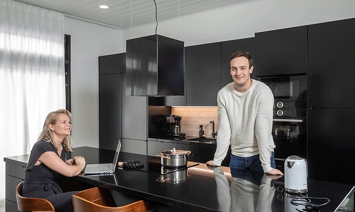 Ett par som trivs i sitt nya svarta Epoq-kök. Kvinnan sitter framför bärbar dator vid köksön och mannen står framför spisen.