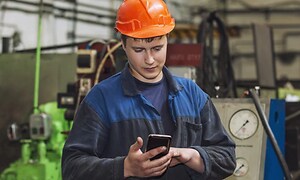 Man i arbetskläder och hjälm står på en byggarbetsplats med en mobiltelefon i handen. 