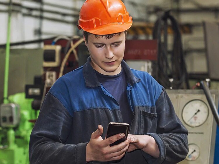 Man i arbetskläder och hjälm står på en byggarbetsplats med en mobiltelefon i handen. 