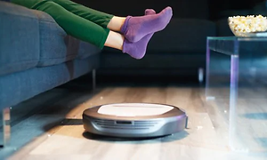 Robotdamsugare städar på golvet medan en person med lila strumpor slappar i soffan. En popcorn skål står på soffbordet.