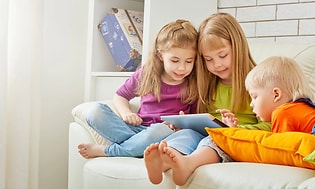 Tre barn, två flickor och en liten pojke sitter tillsammans i en vit soffa och tittar på en surfplatta. 