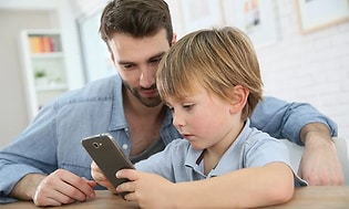 En pojke sitter och tittar på en mobiltelefon medans en vuxen man sitter bredvid och ser vad pojken gör. 