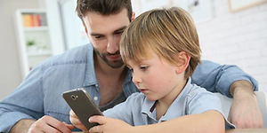 En pojke sitter och tittar på en mobiltelefon medans en vuxen man sitter bredvid och ser vad pojken gör. 
