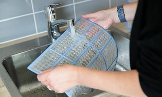 Händer sköljer ett filter från en värmepump under en kran med rinnande vatten. 