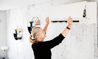 Kvinna öppnar upp locket på en vit värmepump på väggen för att kolla filtret. 
