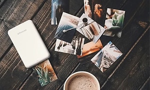 En liten vit portabel skrivare placerad på ett träbord med utskrivna foton som ligger utspritt runt omkring, och en kopp kaffe. 