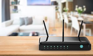 En svart trådlös router med tre antenner, i bakgrunden i oskärpa syns en soffa, TV och ett stort matbord.