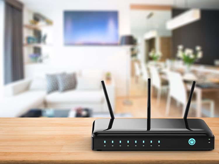 En svart trådlös router med tre antenner, i bakgrunden i oskärpa syns en soffa, TV och ett stort matbord.