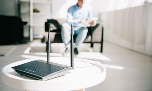 Trådlös router placerad på ett vitt bord, i bakgrunden i oskärpa syns en man som sitter i en fåtölj.