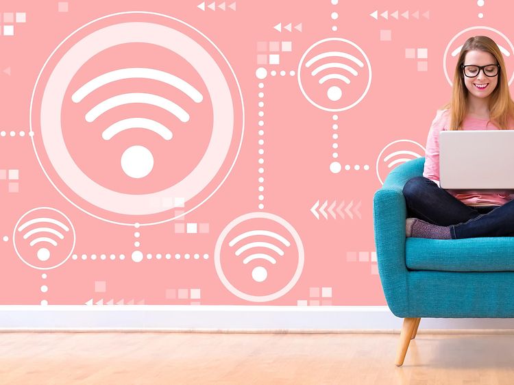 Kvinna sitter med korslagda ben i en turkos fåtölj oc har en laptop i knät. Väggen bakom är rosa med en massa WiFi-symboler.