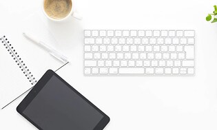 En liten svart surfplatta på ett bord med vitt tangentbord och en vit mus, kaffekopp och anteckningsblock vid sidan.  