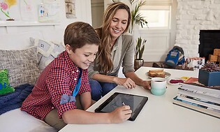 En pojke sitter vid ett bord med en Google Chromebook framför sig, en vuxen kvinna sitter bredvid med en kopp kaffe och ser på. 
